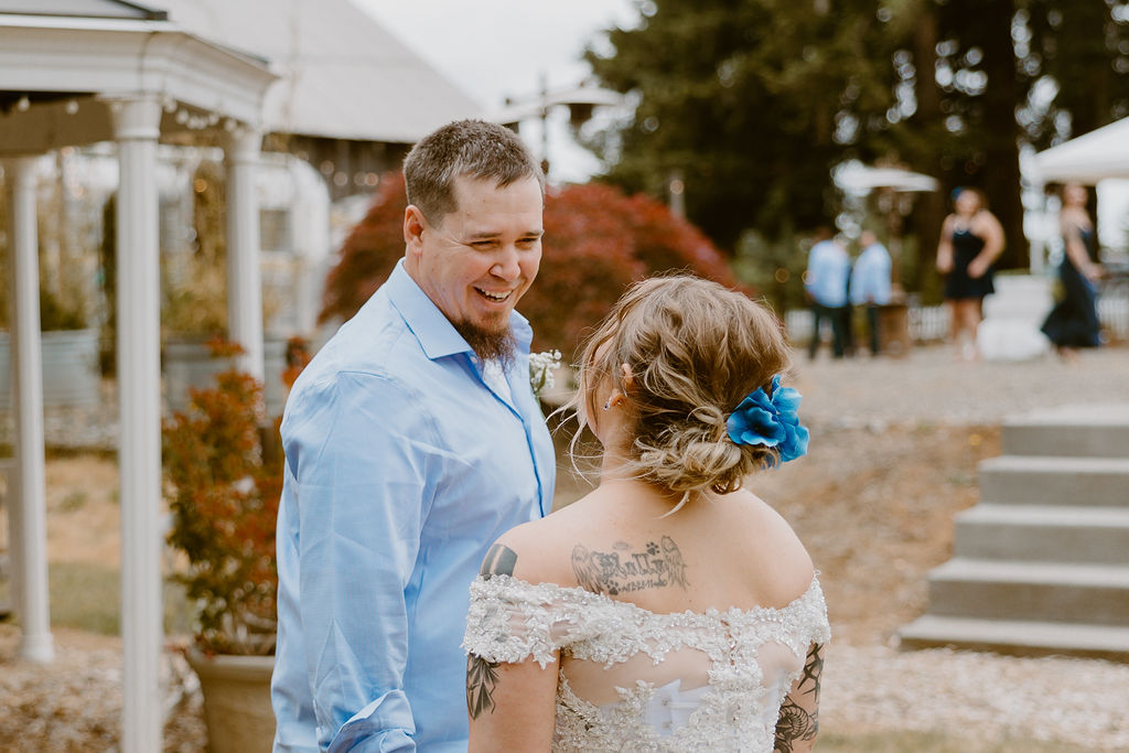 A groom smiles warmly at the bride, sharing a joyful moment outdoors with guests in the background.