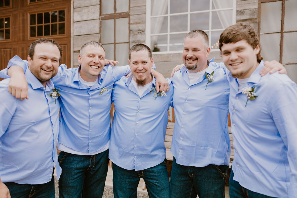 The groom stands with his groomsmen, all smiling and posing in matching light blue shirts.