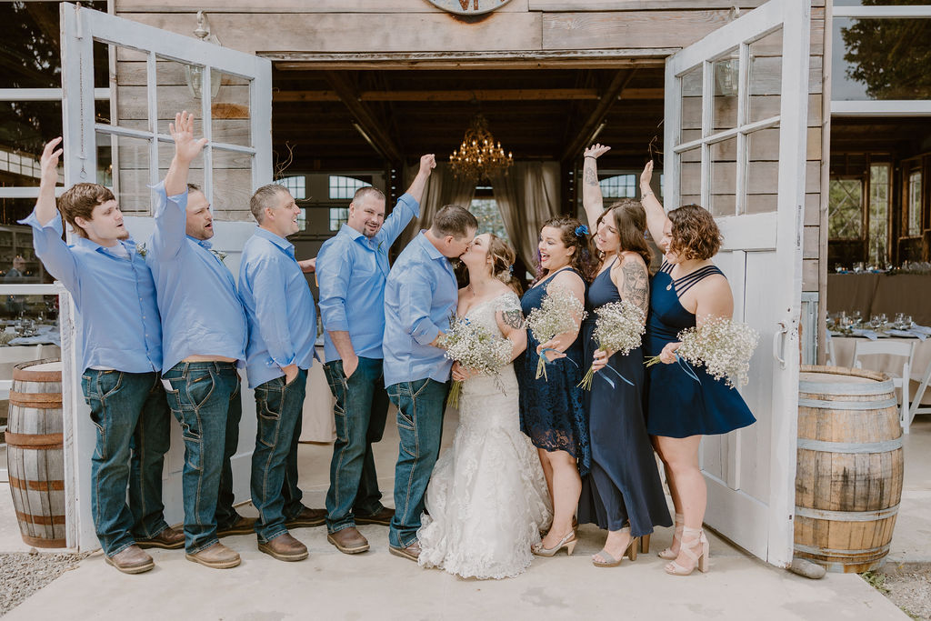 The wedding party celebrates as the bride and groom share a kiss in front of a rustic barn.