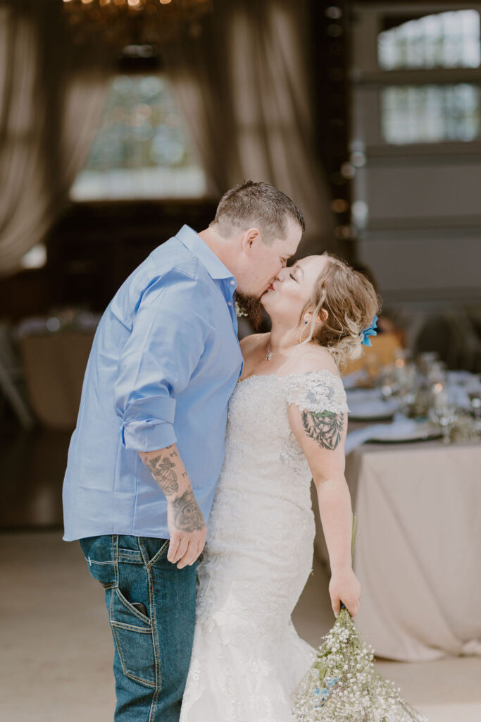A bride and groom share a romantic kiss indoors, highlighting their tattoos.