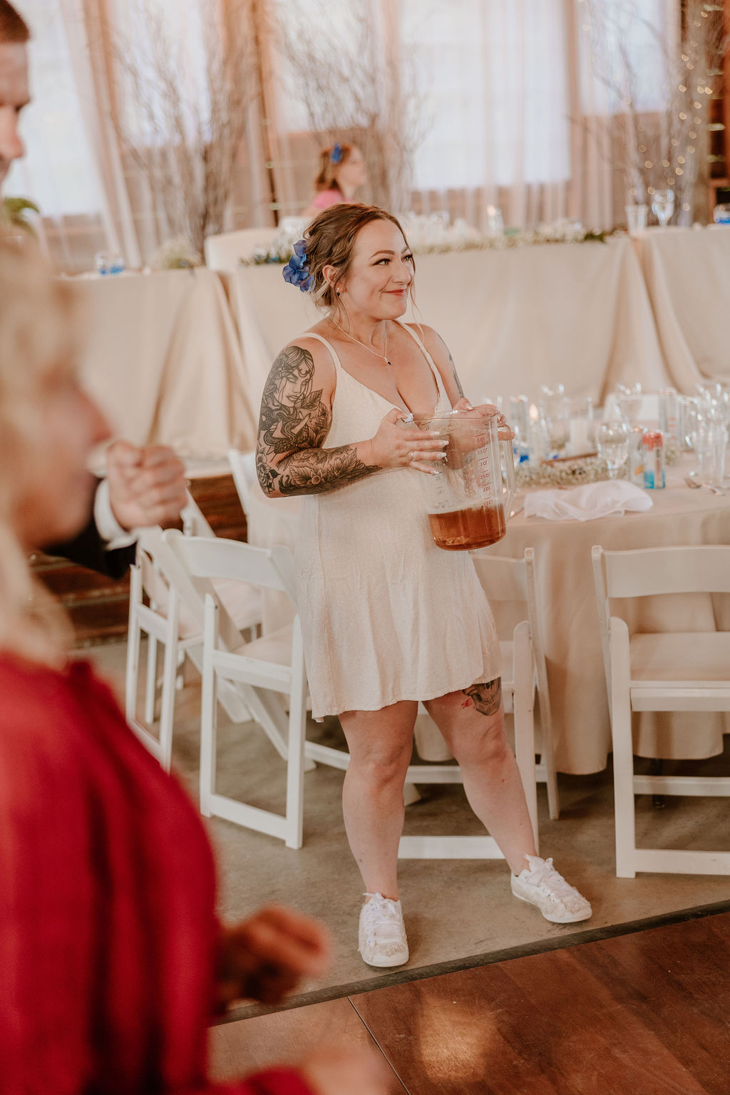 A bride, dressed in a short white dress, holding a large pitcher, smiles during the reception.