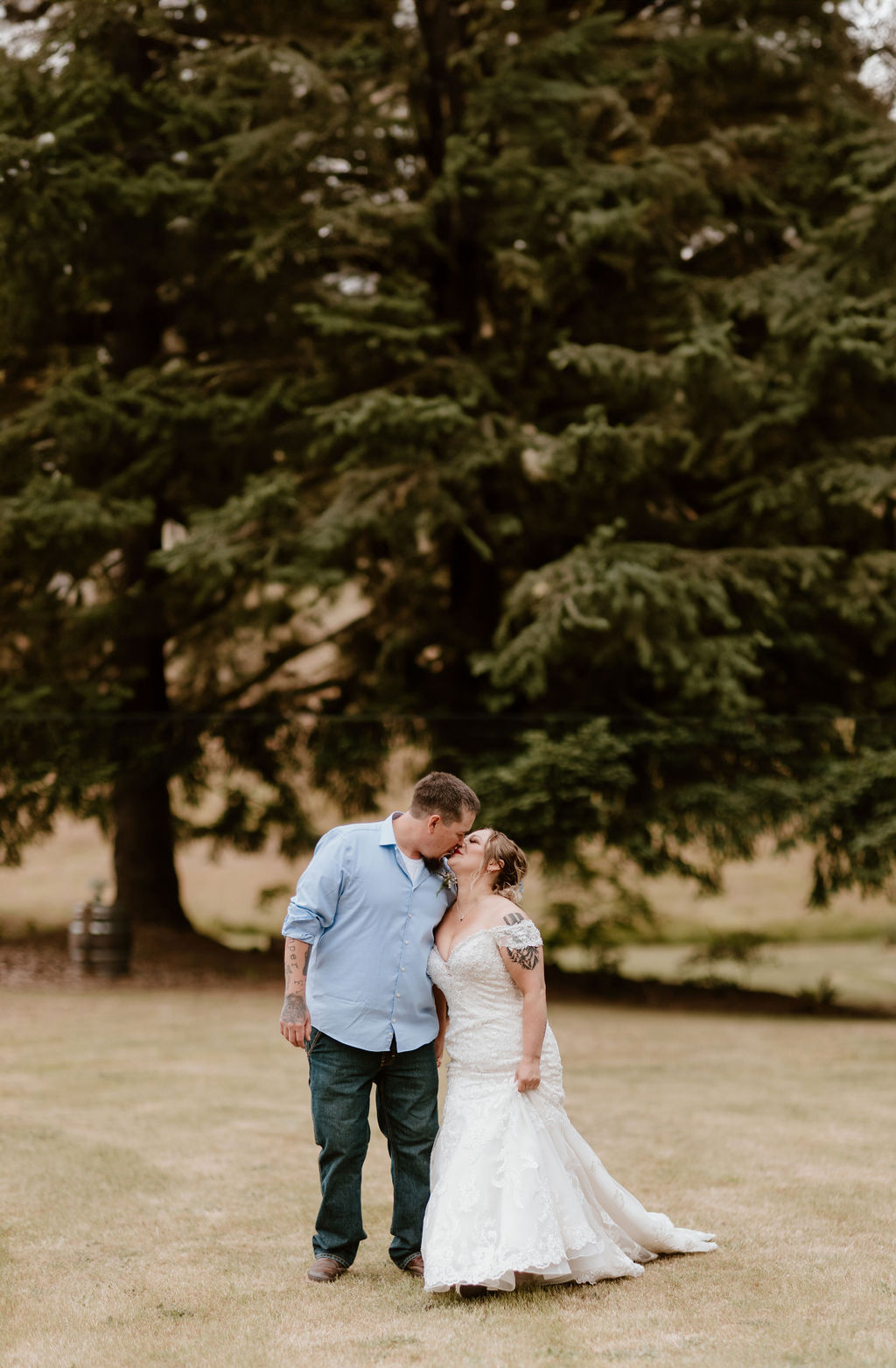A bride and groom share a kiss outdoors in a serene setting with tall evergreen trees.