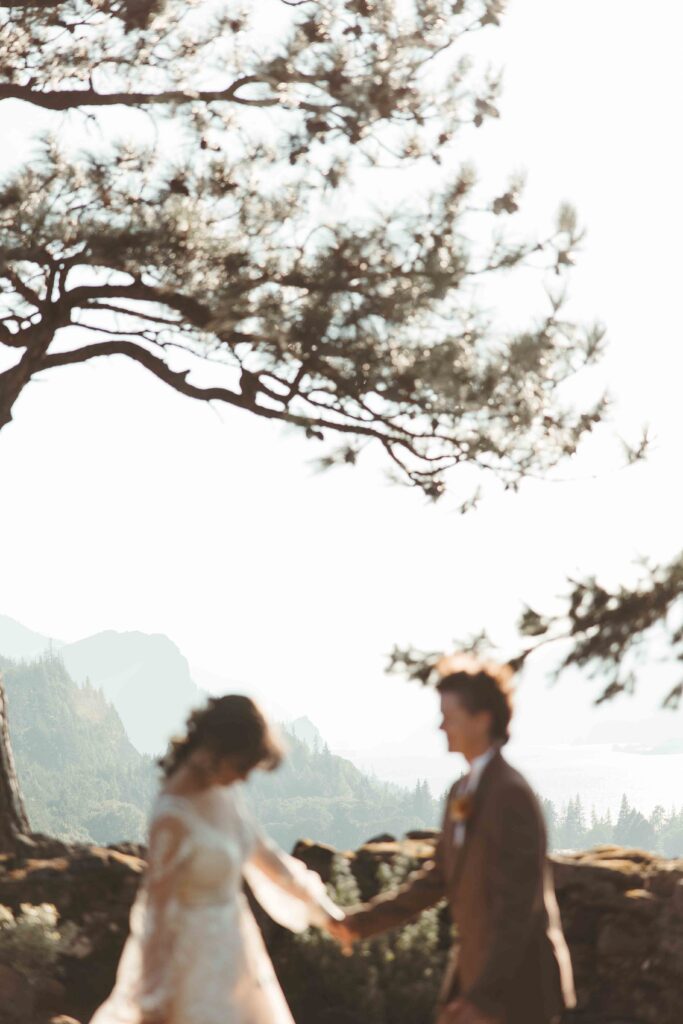 Brides laughing together with sweeping Columbia River Gorge cliffs and evergreen forests in the background.