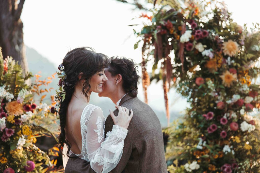 a bride leans in to kiss her partner while they whisper into her ear