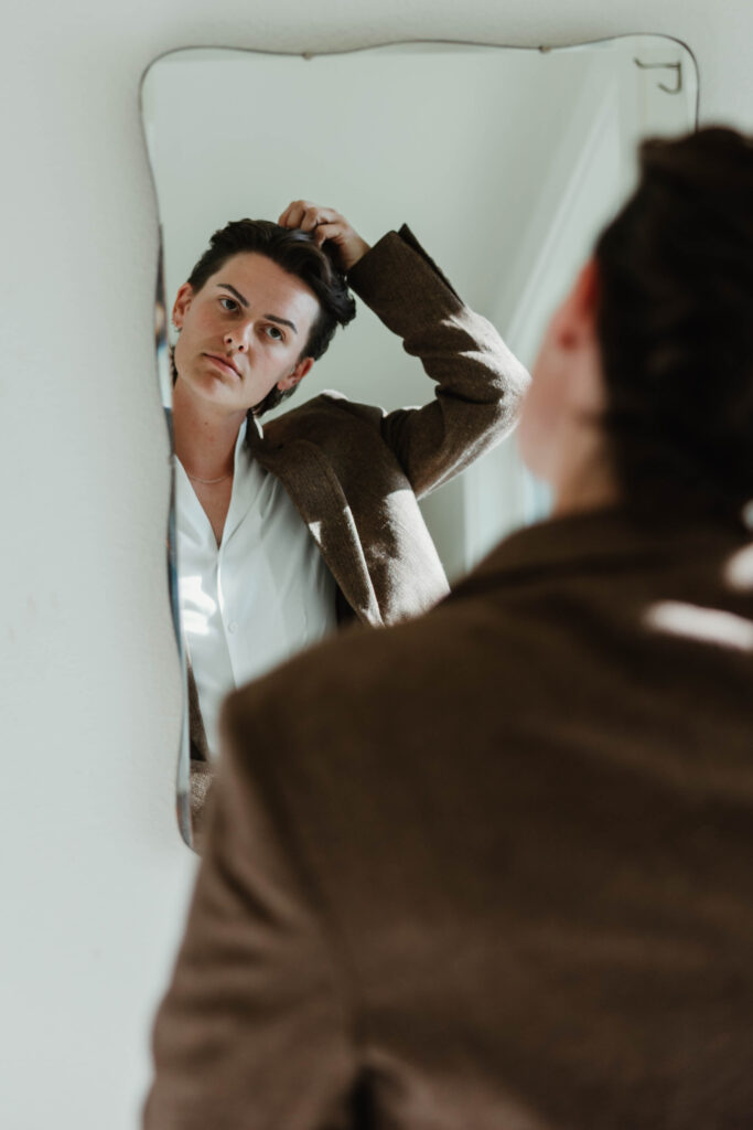 A lgbt bride wears a brown suit and open collar white shirt and puts the finishing touches on their hair for their columbia river gorge elopement