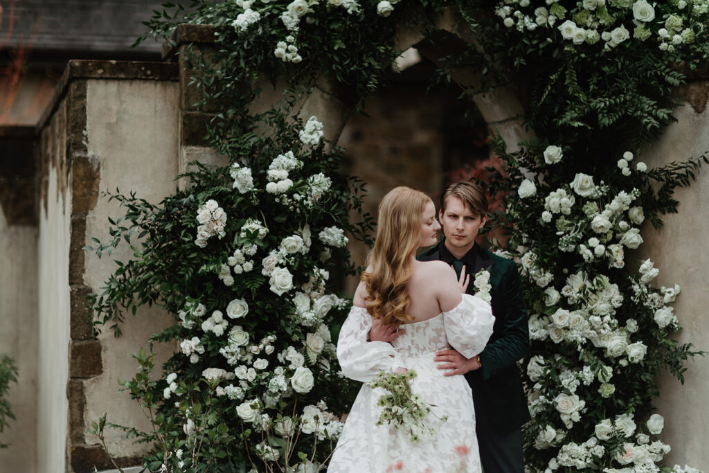 bride and groom pose in front of their wedding arch filled with white roses on their wedding day