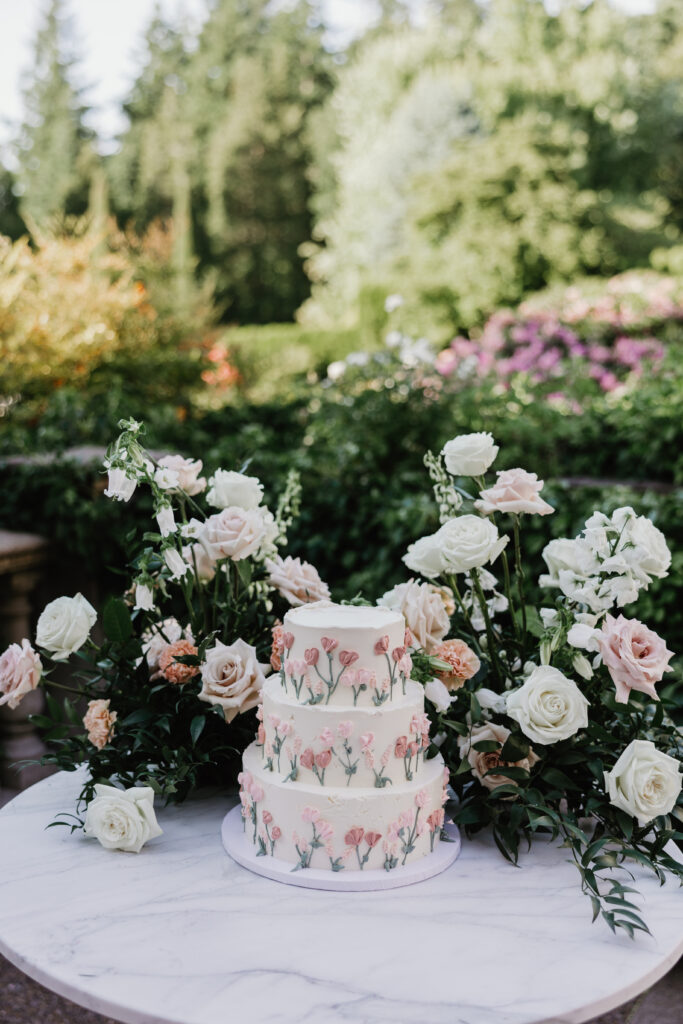 White Floral three tier cake and flower arrangement in a lush green garden during a wedding reception in Vancouver WA