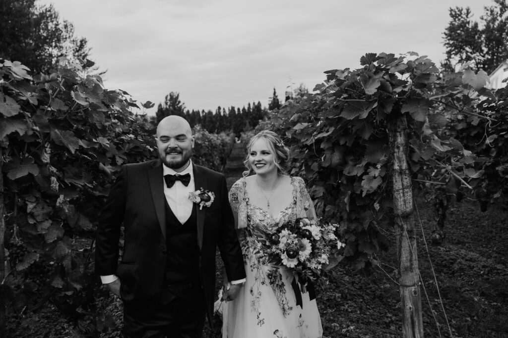 Black and white photo of the bride and groom smiling and walking together through the green vineyard at the end of their wedding day near Vancouver WA