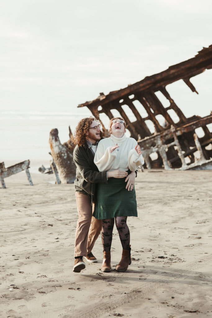 Engaged couple laughing together in front of the wreck of the Peter Iredale in Astoria OR
