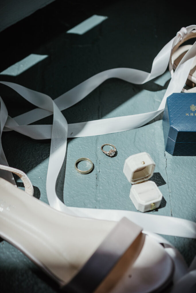 Bridal details flatlay including engagement ring, wedding bands, and invitation suite on a garden surface