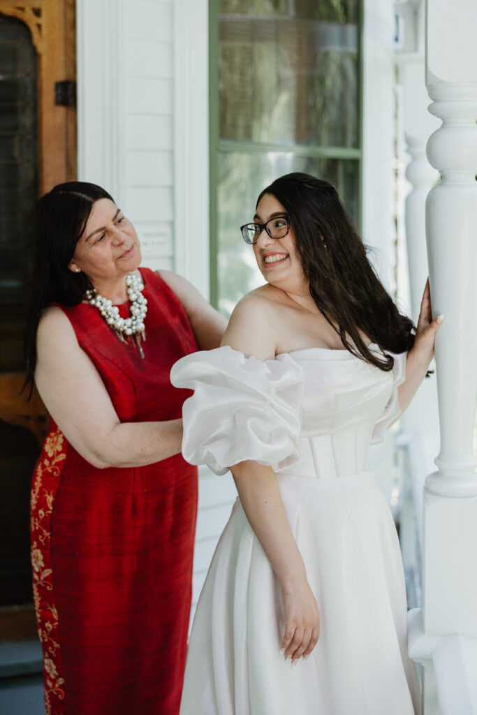 Bride smiling at her mom while they laugh as they zip up the brides white wedding dress