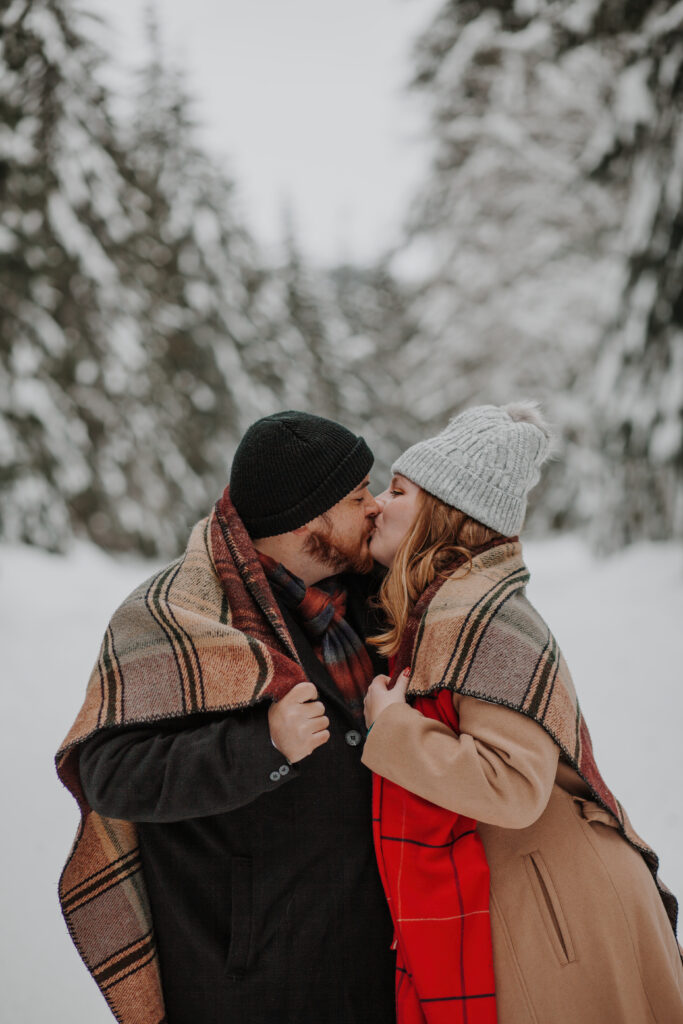 josh and alysa share a sweet kiss under a warm blanket in their engagement session in the snow